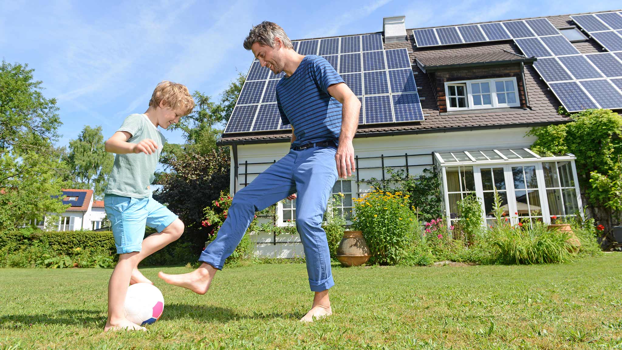 Mann und Kind spielen Fussball auf der Wiese vor einer Solaranlage auf dem Dach.