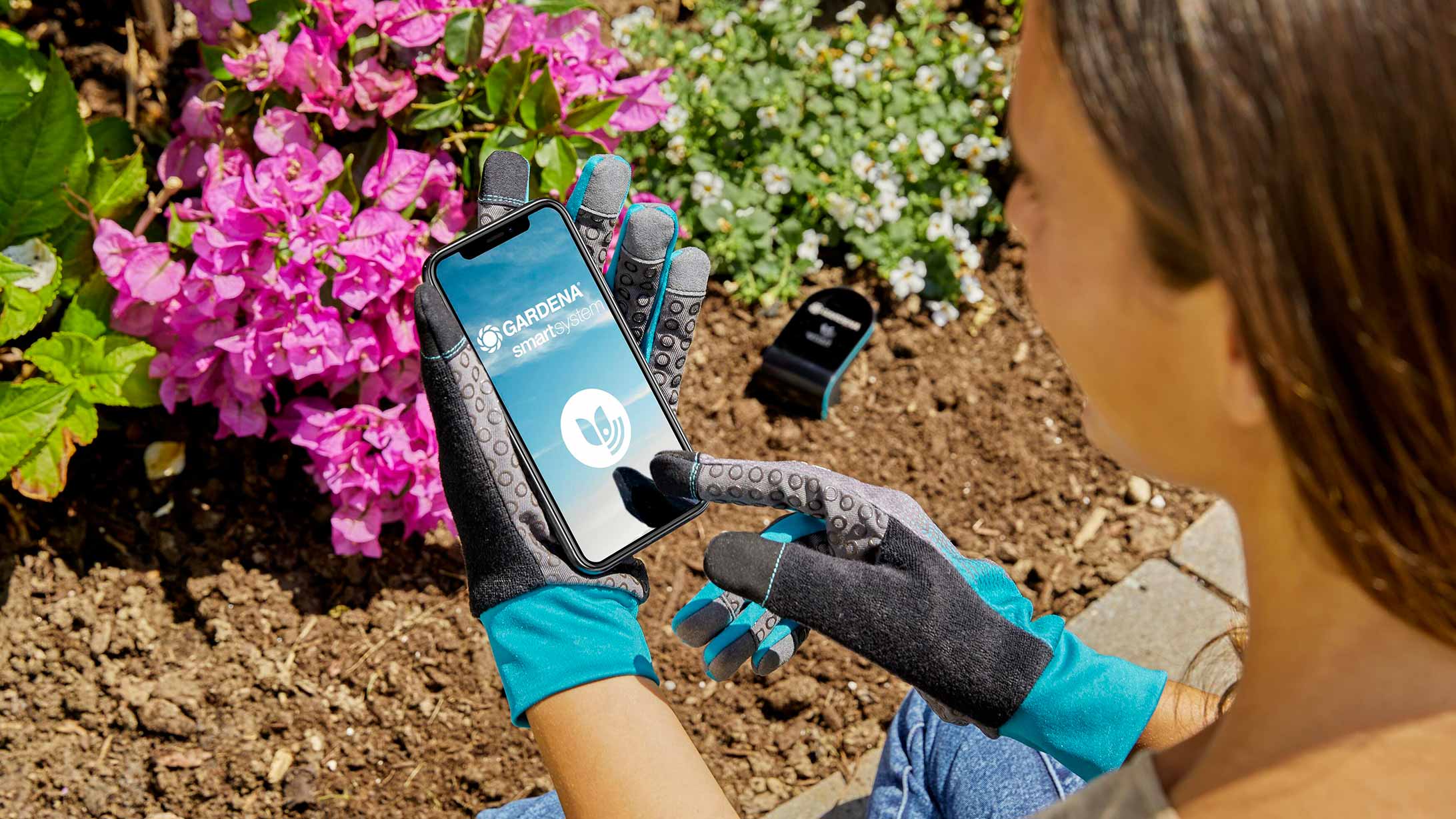Eine Frau bedient mit Gartenhandschuhen ein Smartphone mit der Smart System App von Gardena. Im Hintergrund ein Blumenbeet.
