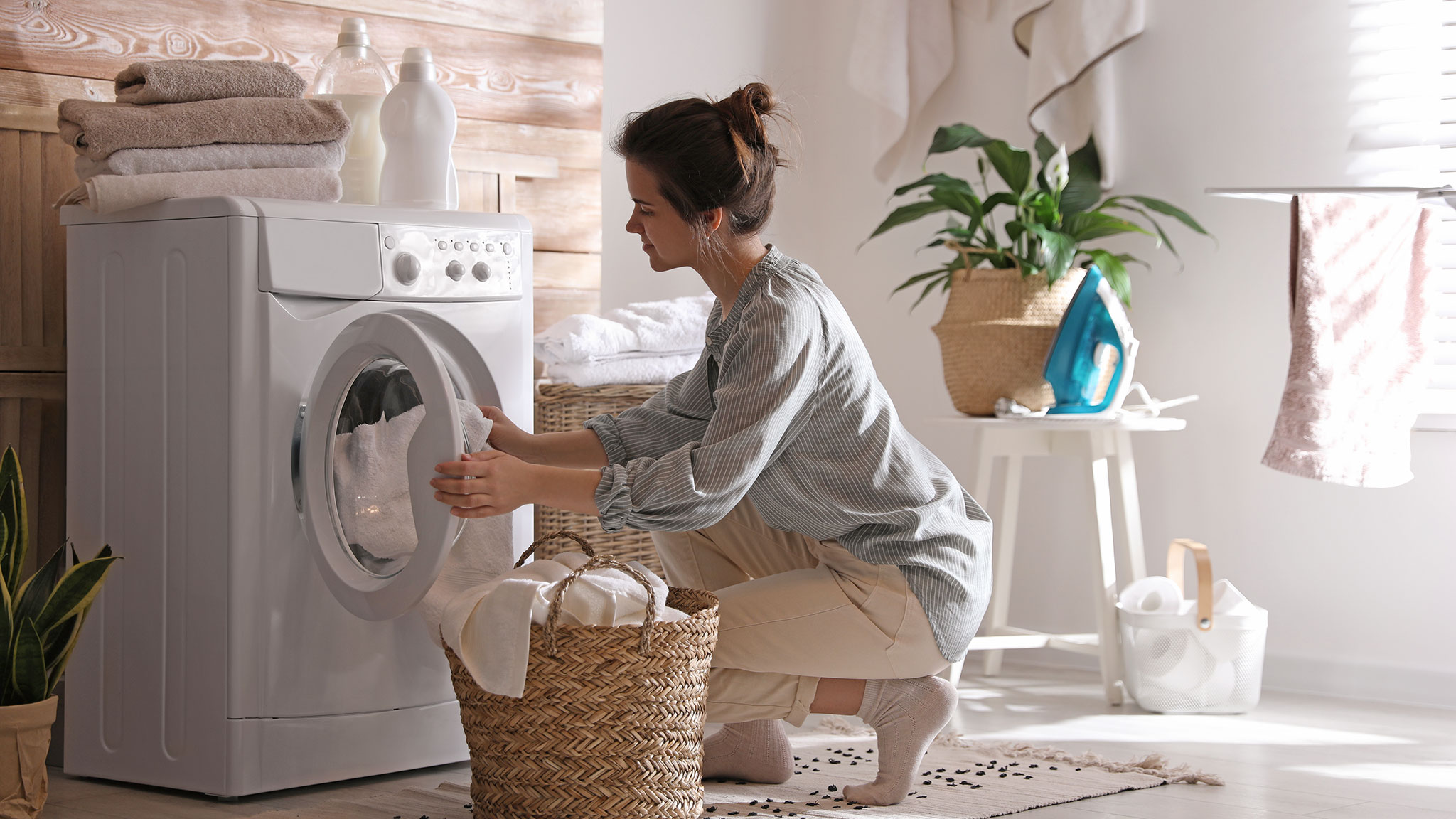 Frau mit Wäschekorb füllt Waschmaschine in Waschraum.