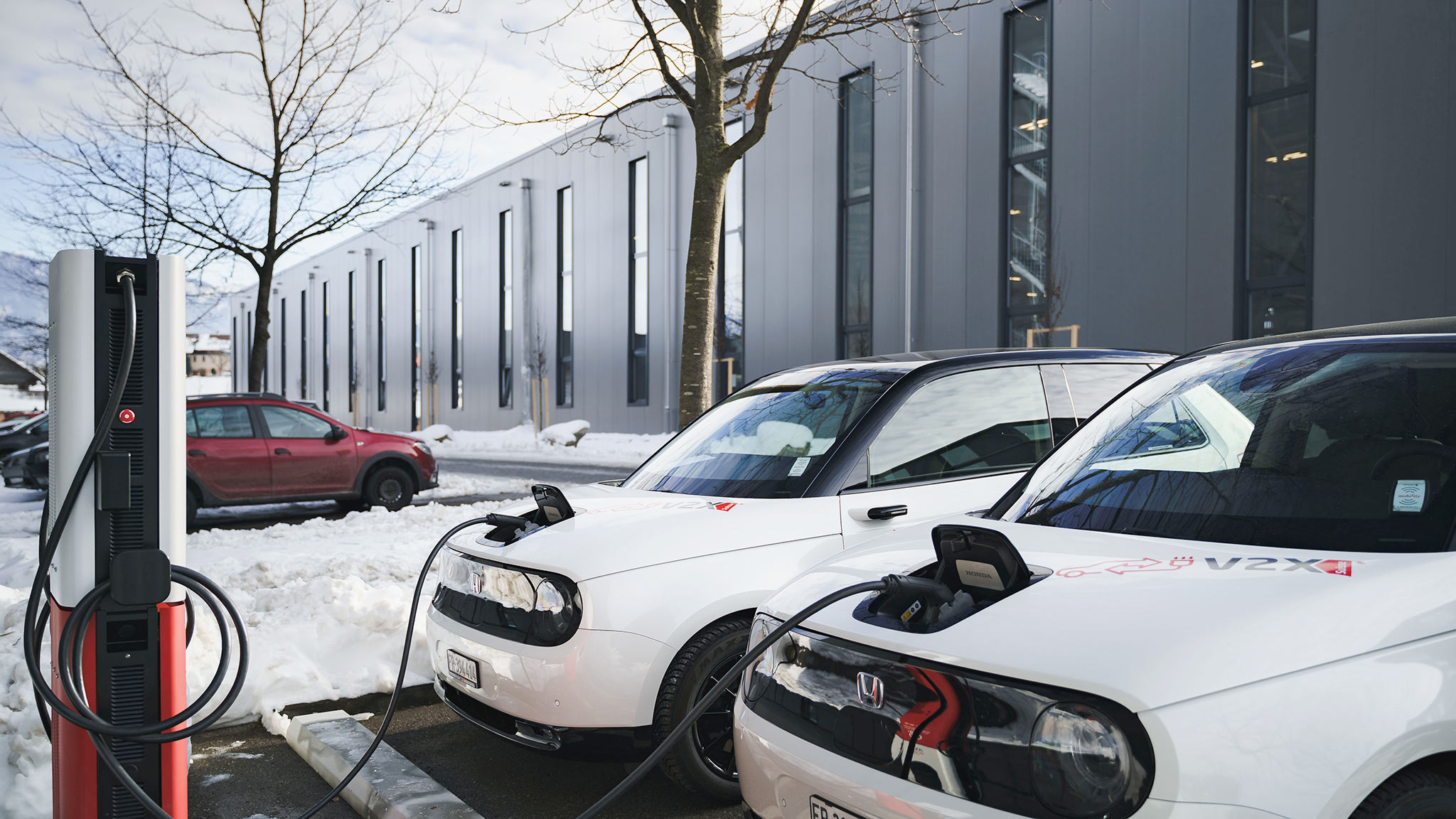 Zwei Elektroautos stehen auf verschneiten einem Parkplatz vor einem Gebäude und werden an einer Ladesäule geladen.